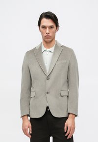 Světle šedý kordjakový blazer s dvěma předními kapsami, dvěma tlačítky a klopovými límci. Nosený přes světle šedou košili na zip.