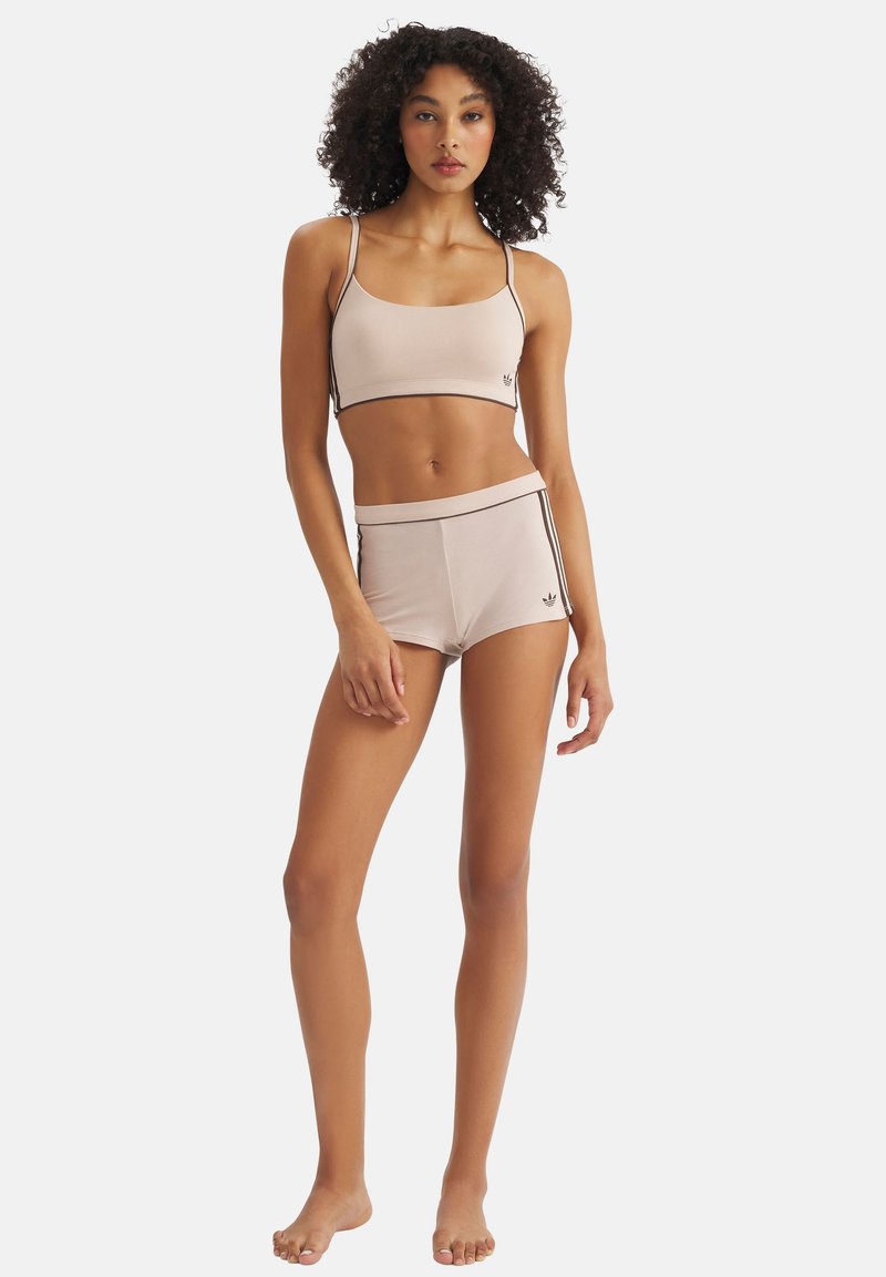 Jeune femme debout, pieds nus, portant un débardeur beige court et un short assorti avec de fines rayures noires sur les côtés.