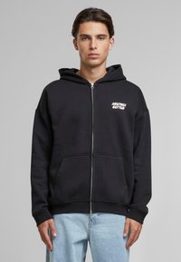 Hanorac negru cu fermoar, confecționat dintr-un material moale, având un imprimeu al logo-ului în față, buzunare tip kangaroo și mansete și tiv din tricot. Asortat cu blugi de culoare bleu deschis.