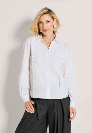 Wit overhemd met knopen, kanten afwerking aan de zijkanten, lange mouwen en een klassieke kraag, gecombineerd met donkere textuurbroeken.