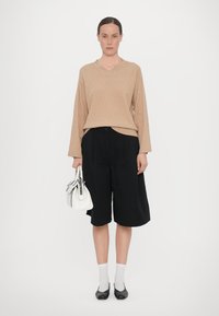 Maglione beige lavorato a maglia con scollo a V e motivo a diamante, abbinato a pantaloni corti neri a pieghe. Borsa bianca e calzini grigi con scarpe nere.