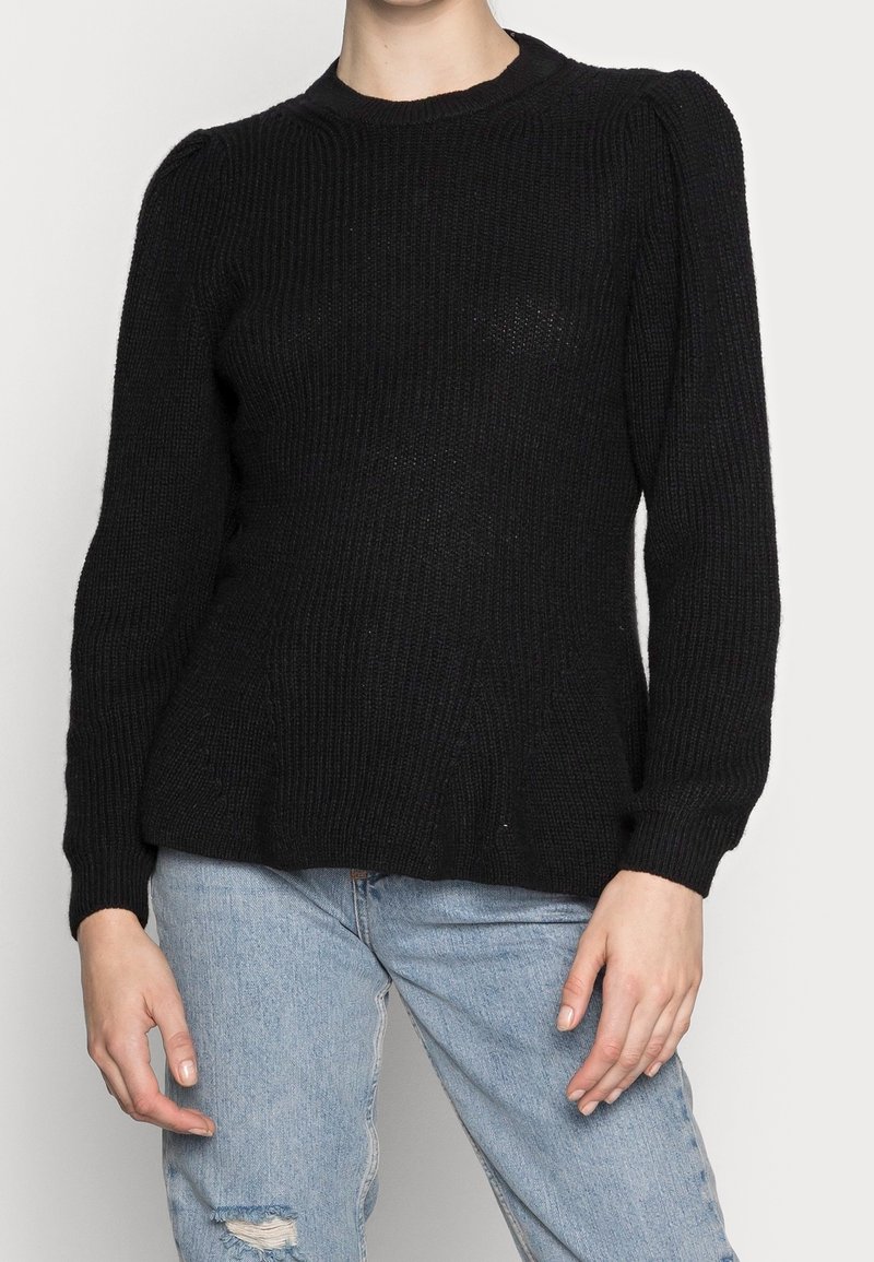 Schwarzer gestrickter Pullover mit Rippstruktur, Rundhalsausschnitt und langen Ärmeln; figurbetont und mit leicht ausgestellt Saum. Getragen mit hellen Jeans.