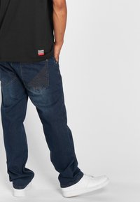 Donkerblauwe denim jeans met een relaxed fit en unieke stiksels op de achterzakken, gecombineerd met witte sneakers. Zwarte t-shirt zichtbaar.