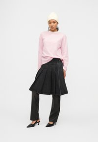 Γυναίκα που φοράει λευκό σκουφάκι, ροζ ριγέ μπλούζα με μακριά μανίκια, μαύρη φούστα με πιέτες πάνω από μαύρο παντελόνι και μαύρα μυτερά τακούνια, στέκεται μπροστά σε λευκό φόντο.