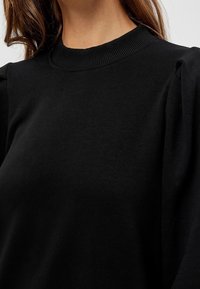 Sweat-shirt noir avec un col côtelé et des manches bouffantes fabriquées dans un tissu doux et texturé. Vue en gros plan mettant en valeur les coutures.