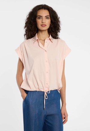 Frau mit lockigem Haar, die eine hellrosa Bluse mit dekorativen Kragen und blaue, hoch taillierte Hose trägt, Hände in den Taschen.