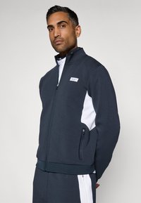 Námornícky zip-up športový kabát s bielymi akcentmi, obsahujúci vysoký golier, bočné vrecká a textúrovanú tkaninu.