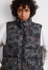 Polstrovaná camouflage vesta s vysokým golierom, zipsom a dvoma prednými vreckami. Farby zahrnujú odtiene šedej, zelenej a čiernej. Viditeľné logo značky.