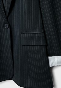 Veste de costume noire à fines rayures avec bouton, poche à rabat et poignet blanc visible à fines rayures.