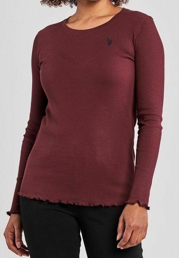 Femme portant un haut ajusté à manches longues en bordeaux à côtes avec des bords festonnés et un pantalon noir, se tenant devant un fond uni.