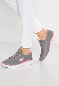 Szare buty sportowe z siateczkową cholewką, różowymi akcentami i białą podeszwą. Posiadają zapięcie na sznurówki i teksturowane powierzchnie.