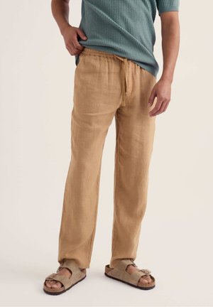 Bărbat purtând pantaloni bej cu șnur, sandale maro deschis și o cămașă scurtă cu mâneci și model dantelat, culoare teal, stând pe un fundal simplu.