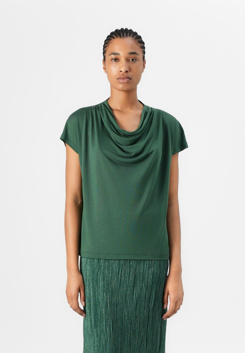 Top verde a maniche corte con scollo drappeggiato; realizzato in un tessuto liscio. Abbinato a una gonna verde plissettata e testurizzata.