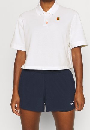 Persoon draagt een wit kort polo-shirt met een klein logo op de borst en marineblauwe Nike-short met een witte swoosh op het been.