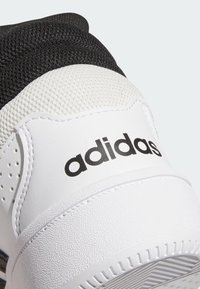 Zbliżenie na piętę białego i czarnego buta sportowego marki Adidas, pokazujące teksturowaną podeszwę i naszywane logo na skórzanym materiale.