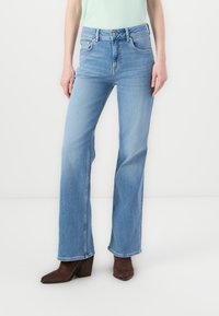 Høyt midjede blå denimjeans med rett ben, lys vask, subtil slitasje og fem-lommers design. Brukt med brune hælboots.