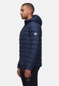 Chaqueta de plumas azul marino con capucha, que presenta un patrón acolchado, bolsillos laterales y un logo en la manga izquierda. Textura suave y brillante.