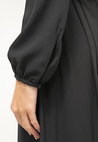 Tissu de robe noire avec une texture lisse, présentant une manche bouffante et un poignet froncé. Main posée sur le côté de la robe.