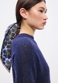 Πλεκτό μπλε πουλόβερ με στρογγυλή λαιμόκοψη. Τα μαλλιά είναι δεμένα με ένα μεταξωτό μαντίλι με σχέδια που περιλαμβάνουν μαύρα σχήματα και πράσινες λεπτομέρειες σε μπλε φόντο.