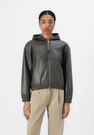 Giacca con cappuccio in pelle verde scuro con zip frontale, polsini ed orlo elasticizzati. Indossata sopra pantaloni beige, caratterizzati da una texture elegante e liscia.
