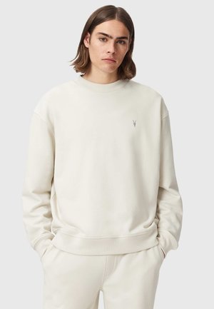 Jong persoon met schouderlang haar, gekleed in een effen gebroken wit sweatshirt en bijpassende broek, handen in de zakken, neutrale achtergrond.