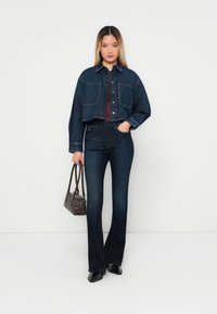 Denim jack met twee voorzakken, donkere was, gecombineerd met bijpassende flared jeans. Model houdt een kleine bruine handtas vast en staat op een neutrale achtergrond.