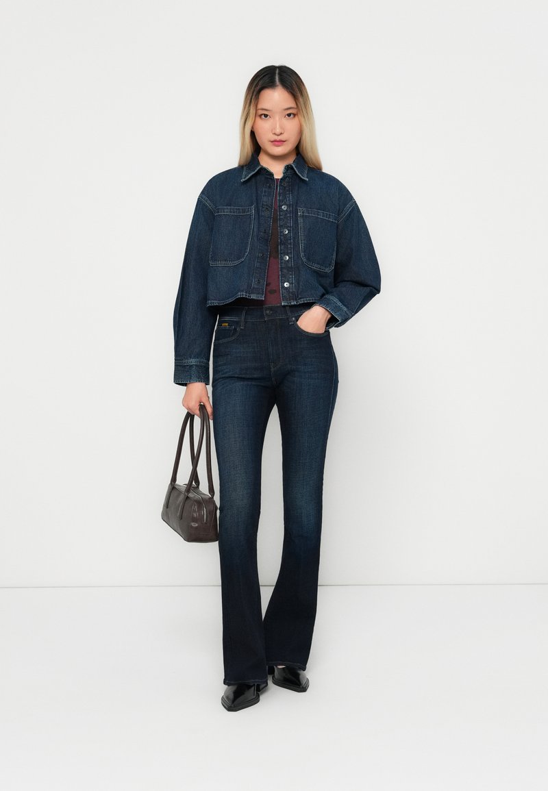 Denimjakke med to forlommer, mørk vask, parret med matchende flagrende jeans. Modellen holder en lille brun håndtaske og står foran en neutral baggrund.