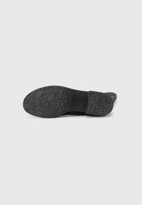 Semelle de chaussure noire avec motif de grip texturé et détail de branding. Le matériau semble être du caoutchouc, avec un design plat et des bords arrondis.