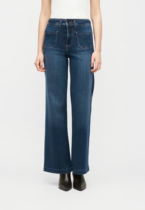 Naine kannab pükse, mis on tumedat sinist värvi, kõrge vöökohaga ja laia säärega, ees patch-taskutega ning mustade terava ninaga saapadega, seistes ühevärvilise tausta ees.