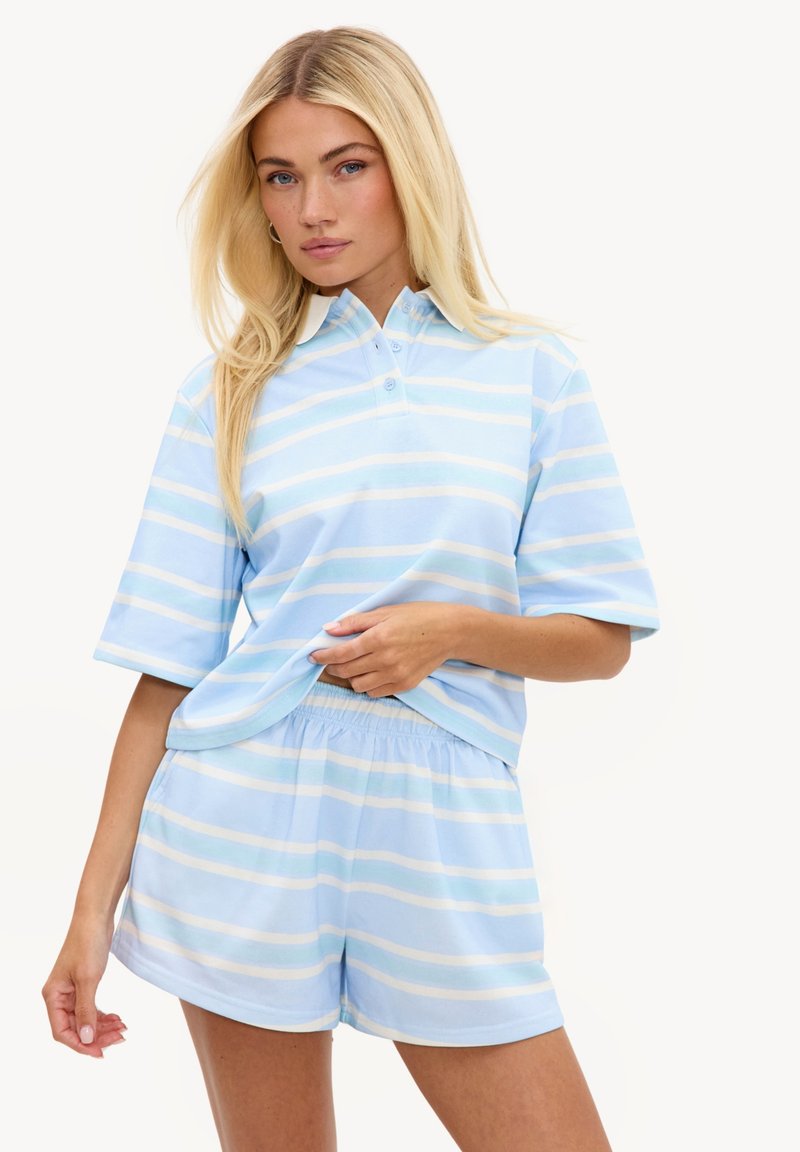 Femme aux longs cheveux blonds portant un polo rayé bleu clair et blanc et un short assorti, debout sur un fond blanc.