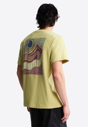 Jasnożółty bawełniany T-shirt z grafiką przedstawiającą góry i fale w fioletowym i niebieskim kolorze, a pod spodem napis "THE NORTH FACE".