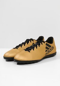 Kultaiset synteettiset jalkapallokengät, joissa on teksturoitu pinta, mustat nauhat ja huomattava musta logo sivussa. Suunniteltu tekonurmipinnoille.