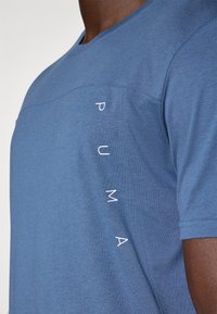 Camiseta corta de manga azul hecha de una mezcla de algodón, con una sutil textura y el logo de "PUMA" impreso verticalmente en el lado izquierdo.