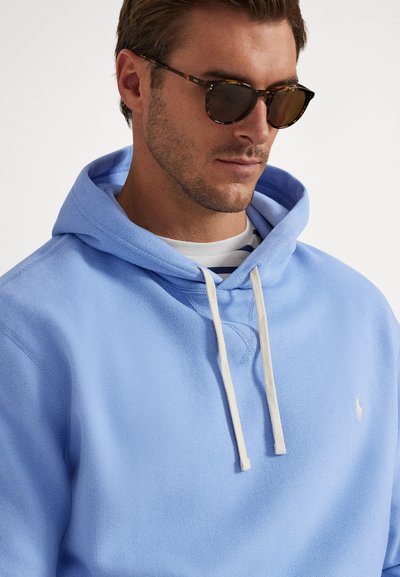 Šviesiai mėlynas džemperis su gobtuvu, pagamintas iš minkštos medžiagos, turi raištelį prie gobtuvo, lowonuotus rankogalius ir mažą logotipą krūtinės srityje. Dėvimas virš dryžuoto marškinėlio.