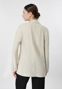 Femme aux cheveux foncés coiffés en chignon bas, portant un blazer beige ample sur un pantalon noir, vue de profil devant un fond blanc neutre.