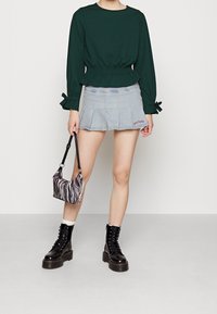 Grünes elastisches Taillenshirt, hellblaue Jeans-Falten-Minirock mit rotem Logo, schwarze Schnür-Stiefel und eine gestreifte Handtasche.