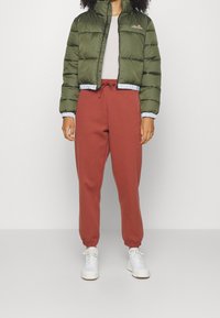 Giacca imbottita verde oliva con colletto alto, rifiniture azzurro chiaro, chiusura con zip; pantaloni jogger color ruggine con polsini elasticizzati; sneakers bianche.