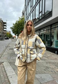 Jachetă matlasată în nuanțe de bej, gri și galben, cu închidere prin butoni și două buzunare. Asortată cu pantaloni lejeri bej. Fundal urban de stradă.