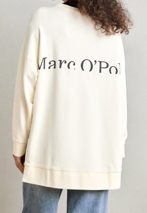 Frau trägt einen übergroßen, cremefarbenen Pullover mit dem Schriftzug "Marc O’Pol" auf dem oberen Rücken, kombiniert mit hellblauen Jeans.