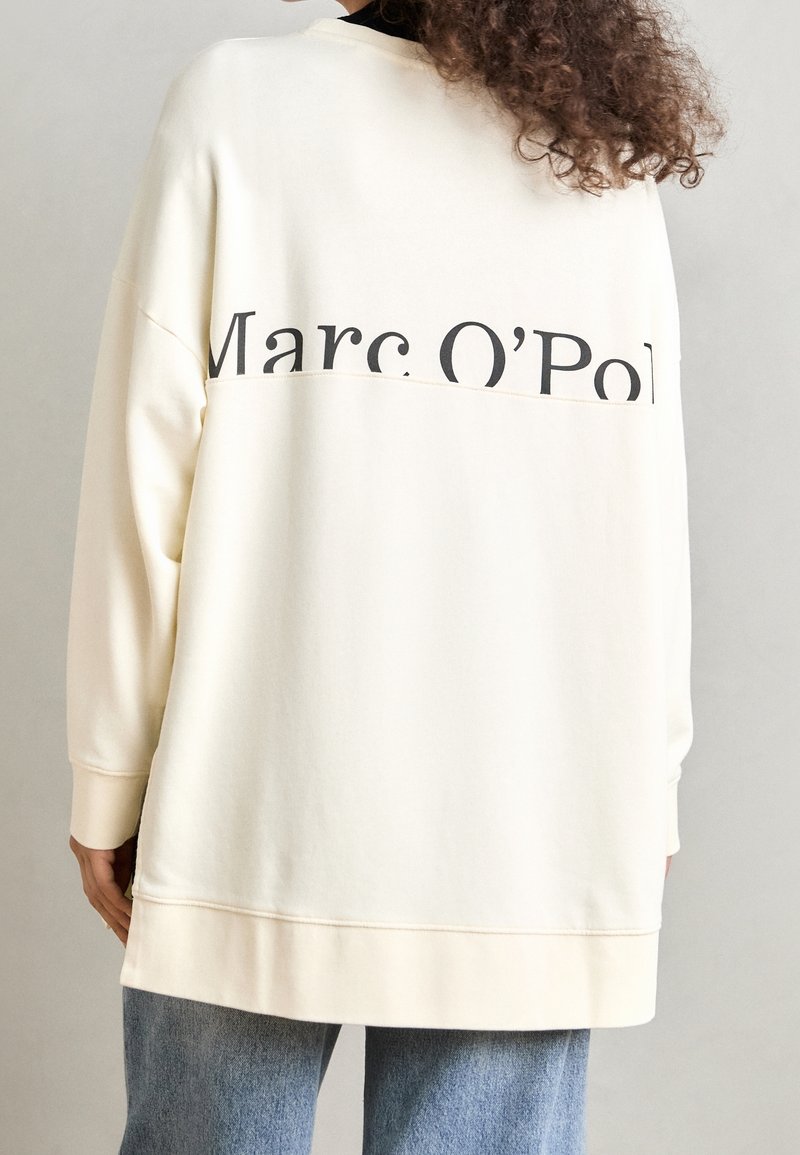 Femme portant un sweat-shirt oversize écru avec l'inscription "Marc O’Pol" sur le haut du dos, associé à un jean bleu clair.
