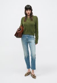 Πράσινη πλεκτή ζακέτα, μπλε ξεβαμμένο τζιν και παπούτσια με λεοπάρ σχέδιο. Κρατά ένα καφέ δερμάτινο τσαντάκι. Απλή, άνετη εφαρμογή με καθαρές υφές.