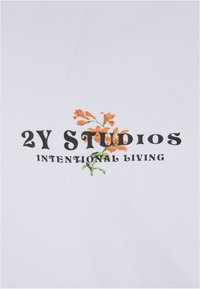 Tricou alb din bumbac cu textul "2Y Studios" și "INTENTIONAL LIVING" într-o fontă neagră îndrăzneață, accentuat cu broderie florală portocalie.