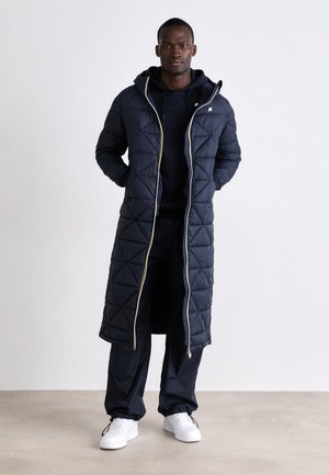 Lange gewatteerde marineblauwe jas met een gouden rits, capuchon en ruitpatroon; gecombineerd met een marineblauwe hoodie en broek, en witte sneakers.