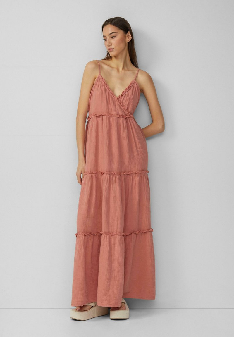 Maxikleid aus weichem terracottafarbenem Stoff, mit dünnen Trägern, einem tiefen V-Ausschnitt, gestuftem Schnitt und Rüschendetails an den Nähten. Plattformschuhe darunter.