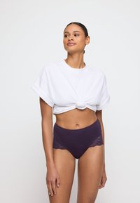 Witte geknoopte t-shirt met opgerolde mouwen, gecombineerd met hooggezette paarse onderbroek met kantaccenten aan de zijkanten.