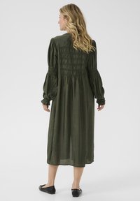 Abito verde oliva a maniche lunghe con schiena arricciata e design fluido. Tessuto liscio e leggero; lunghezza fino al ginocchio. Scarpe nere basse.