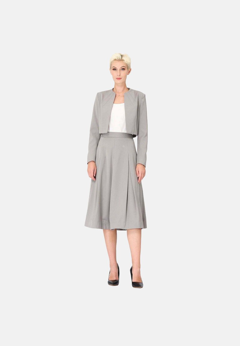 Ensemble deux pièces gris comprenant une veste courte et une jupe plissée arrivant au genou. Tissu lisse avec des manches longues, associé à des talons noirs.