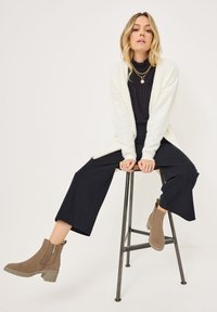 Bottines beiges avec des zippers latéraux, cardigan blanc doux et haut noir associés à un pantalon noir à jambes larges, assise sur un tabouret en métal.