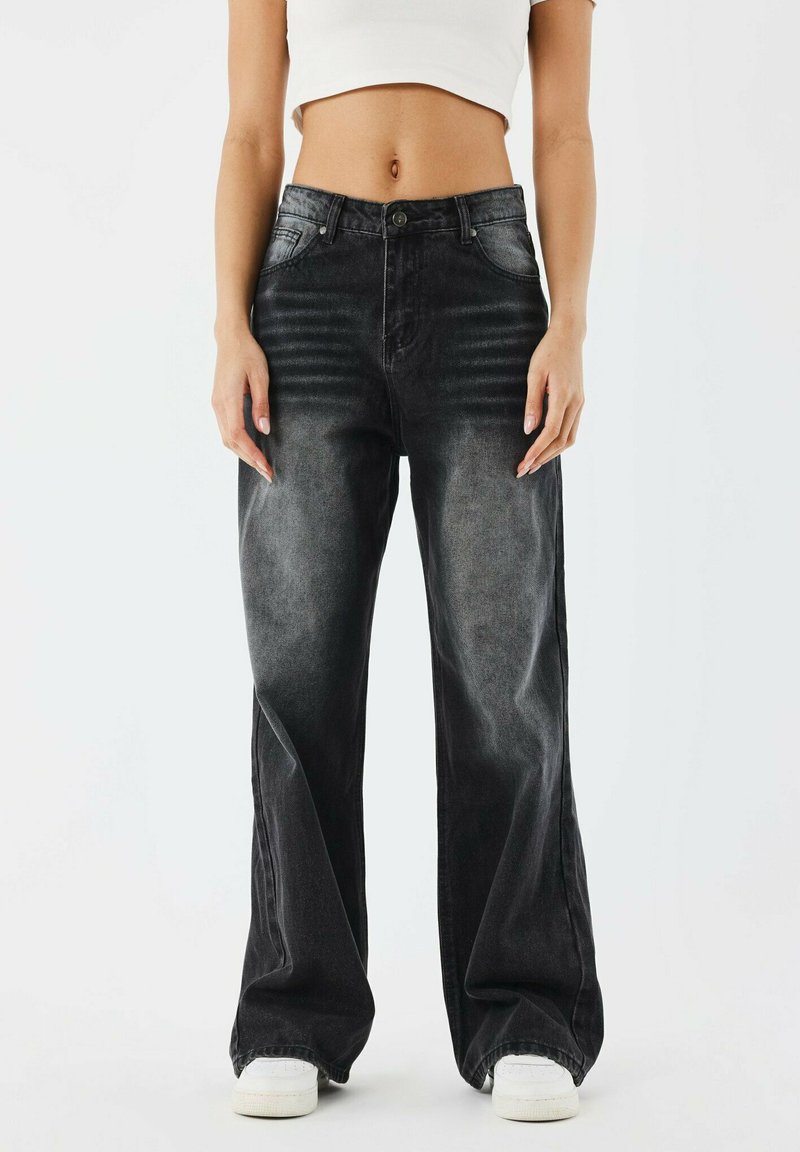 Jean en denim noir à jambes larges avec un délavage, présentant un design classique à cinq poches et un léger effet de froissement à l'avant. Baskets blanches en dessous.