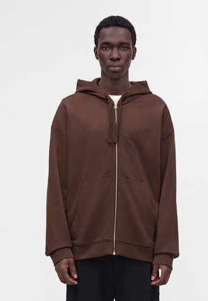 Jeune homme portant un sweat à capuche marron zippé avec des poches avant, debout devant un fond blanc uni.